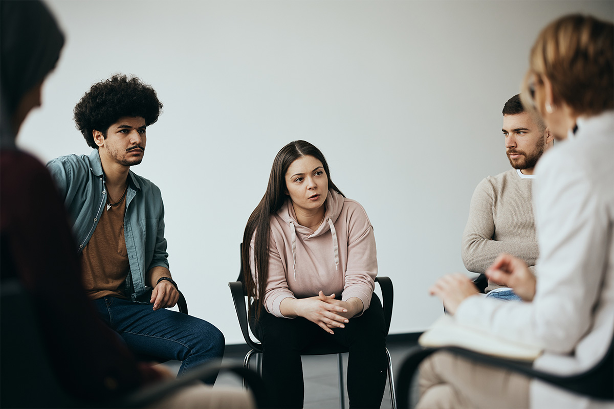 Verschiedene Personen sitzen in einem Kreis und hören einer Frau zu.